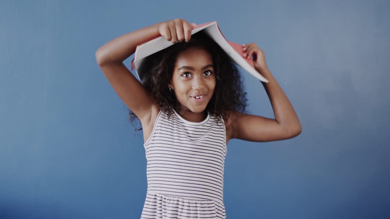 feliz niña afroamericana sosteniendo un libro en la cabeza sobre un fondo azul, cámara lenta
