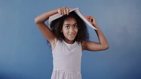 happy african american girl holding book on head on blue background, slow motion