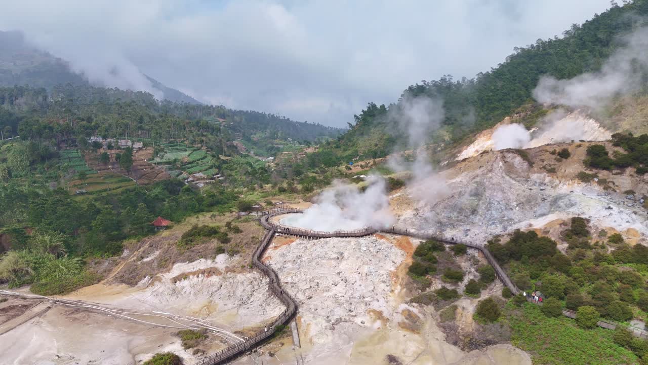 Aerial shot of a volcanic crater emitting steam and gas in Sikidang, Dieng Plateau, featuring tourist walkways and natural geothermal activity