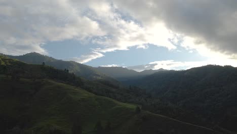 Drone-video-ascending-up-a-mountain-during-sunrise-with-some-light-rays-shining-through-the-mountain-peaks-revealing-the-green-landscape