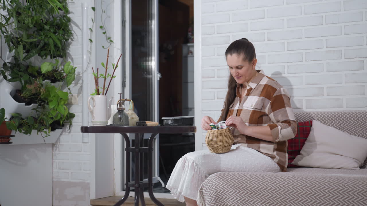 Plant enthusiast seated on sofa near round table selecting card from small wooden basket on lap surrounded by lush green plants white brick wall backdrop cozy indoor gardening activity
