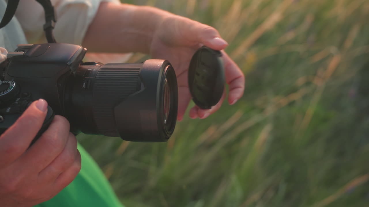 Close up of photographer hands removing lens cover from camera in soft evening light while standing in grassy field wearing green dress, sunlight casting warm glow over natural outdoor setting