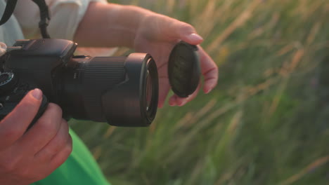 close up of photographer hands removing lens cover from camera in soft evening light while standing in grassy field wearing green dress, sunlight casting warm glow over natural outdoor setting