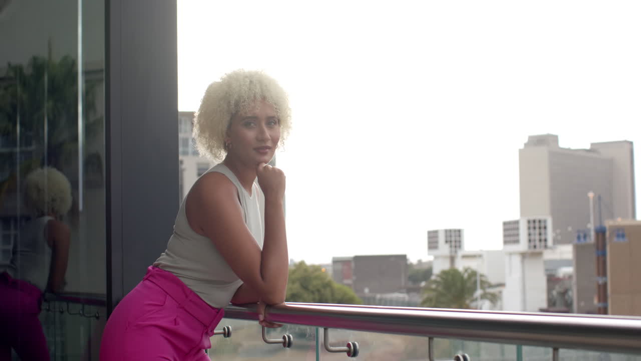 Leaning on balcony railing, woman in pink pants looking at camera confidently, copy space