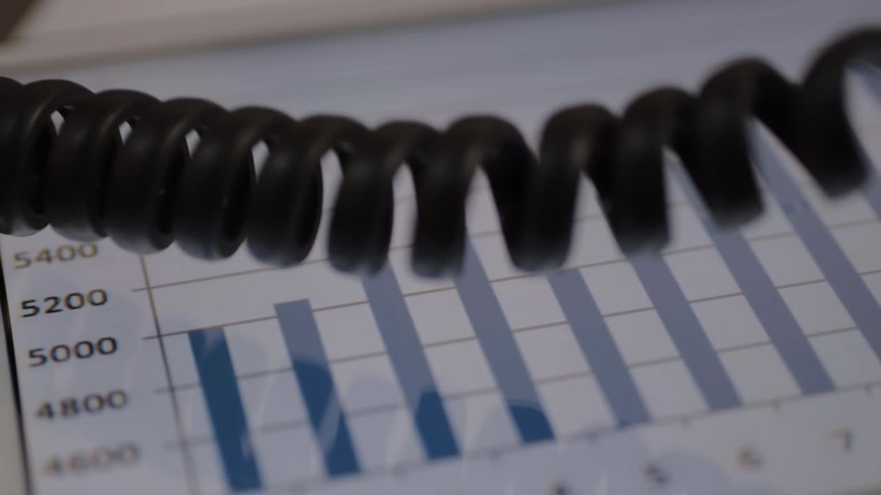 Office phone coil hangs over blue sales chart graph on tablet