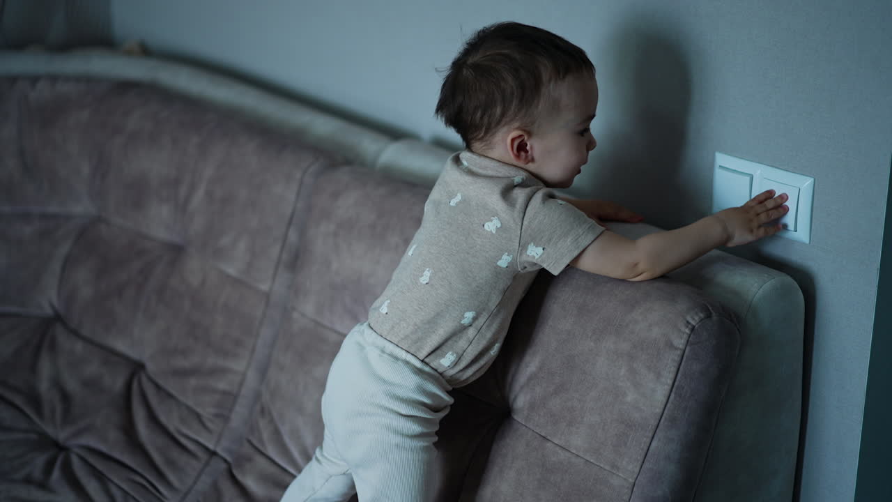Cheerful baby boy stands up and switches on the light in the room. Kid points up happily.