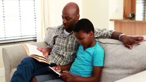 Father-reading-book-and-son-using-digital-tablet-in-living-room-4k-