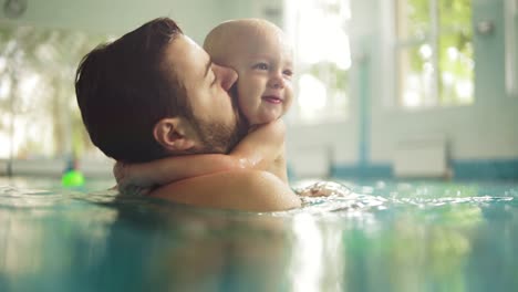 Lindo-Bebé-Y-Su-Padre-Tomando-Clases-De-Natación-En-La-Piscina.-El-Padre-Sostiene-A-Su-Hijo-En-Sus-Manos-Y-Lo-Abraza