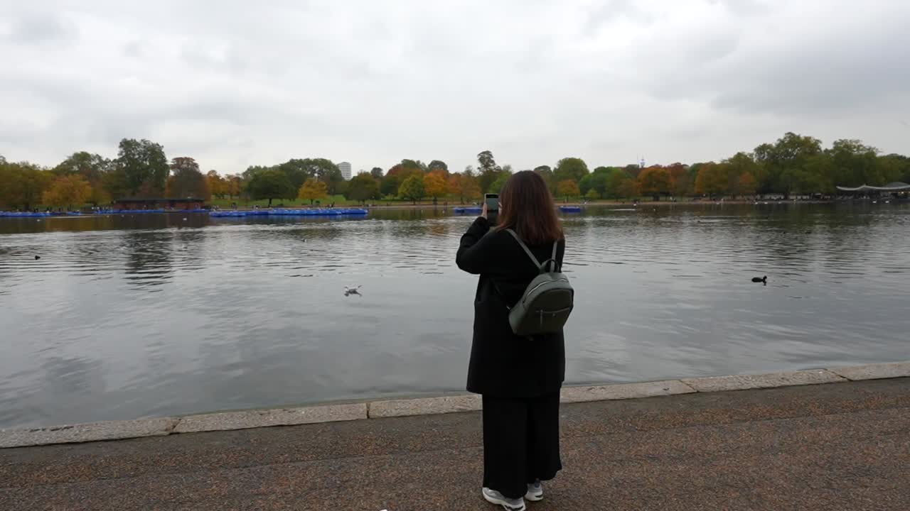 Woman records Hyde Park lake on her phone, capturing the serene view on a cloudy autumn day. London, UK