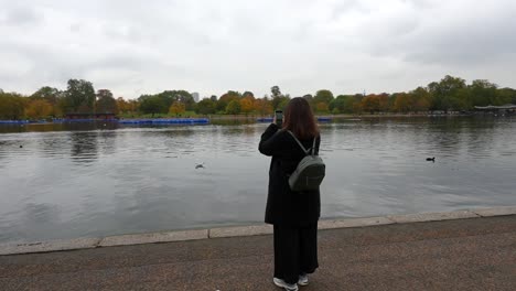 woman records hyde park lake on her phone, capturing the serene view on a cloudy autumn day. london, uk