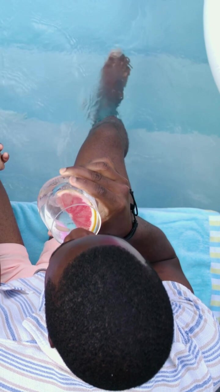 Vertical video: Partner dipping feet in pool, man in stripes stirring, sipping citrus glass to cool