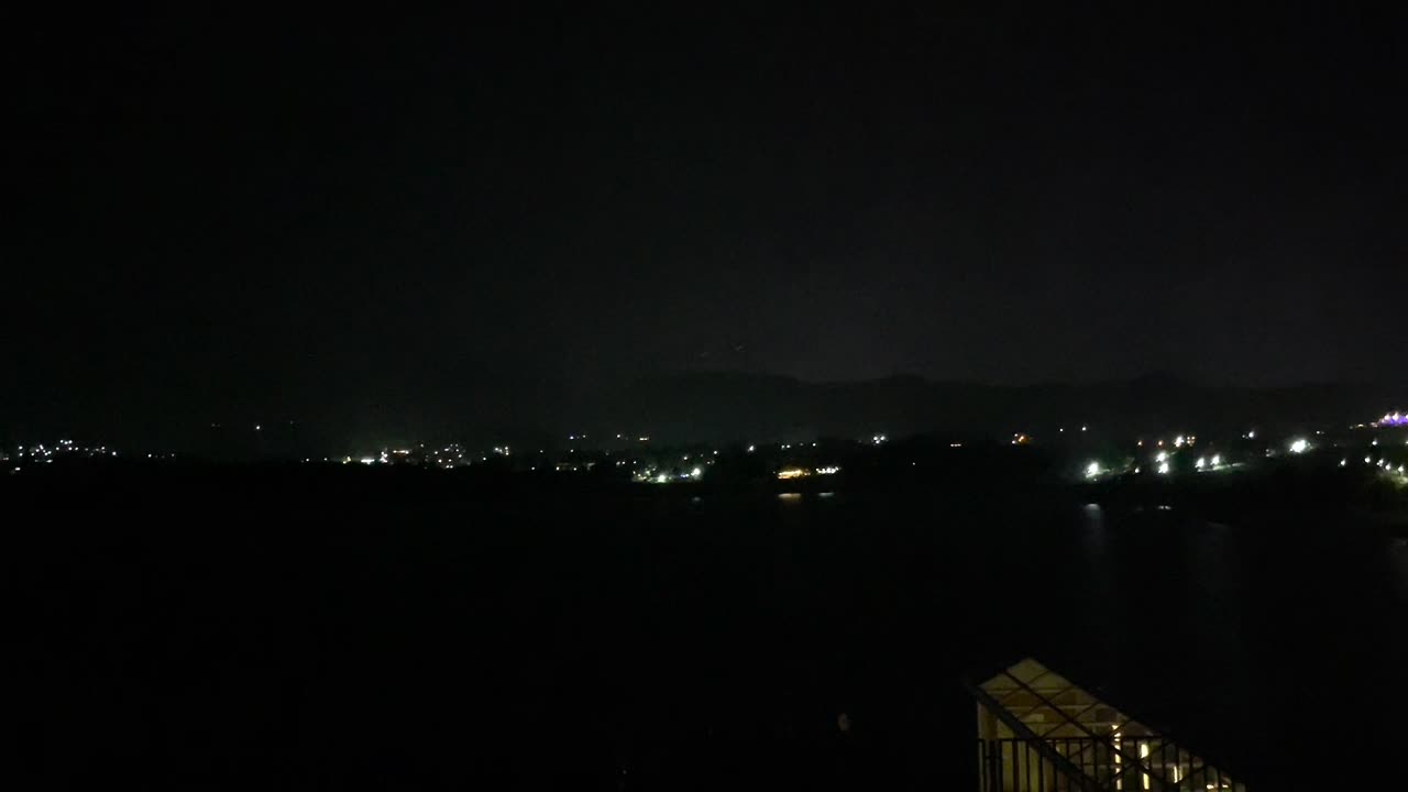 Sky lights up with bright lightning over distant hills and city during dark stormy night