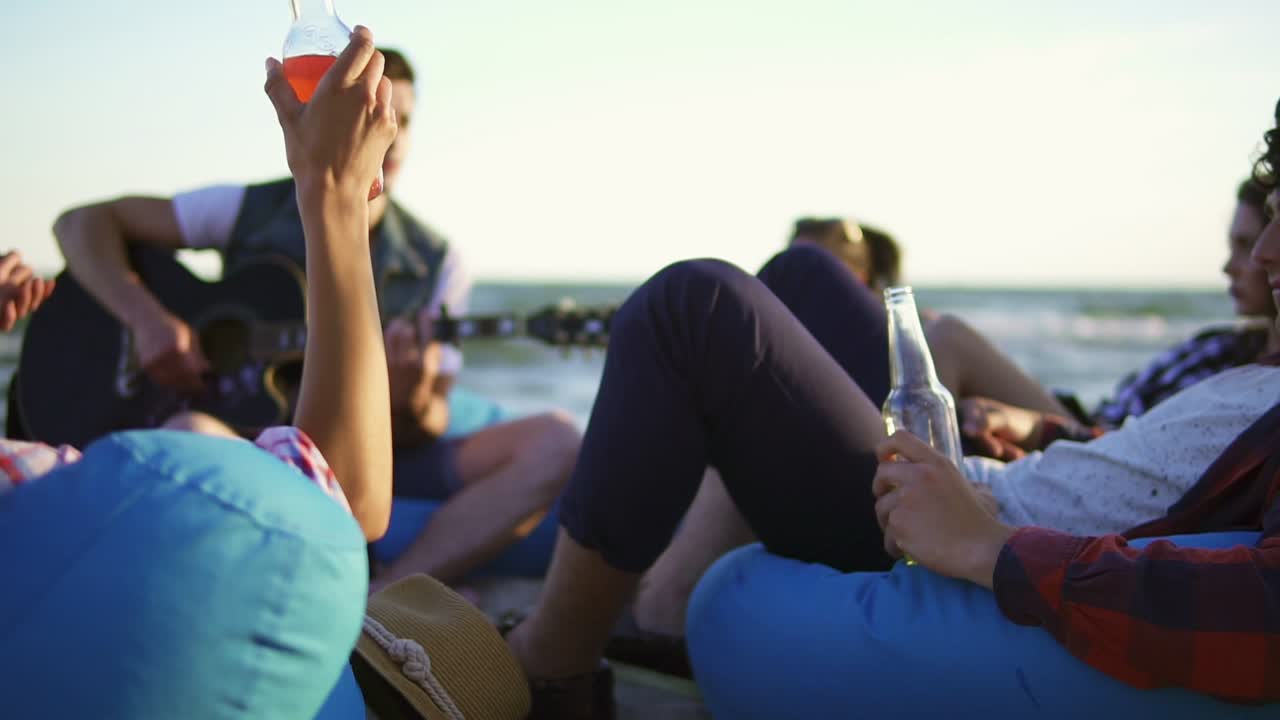 한 무리의 친구들이  ⁇ 테일과 맥주를 마시고 해변의 편안한 의자에 앉아서 해가 지는 여름 저녁에 기타를 연주하는 친구를 듣고 있습니다. 느린 촬영