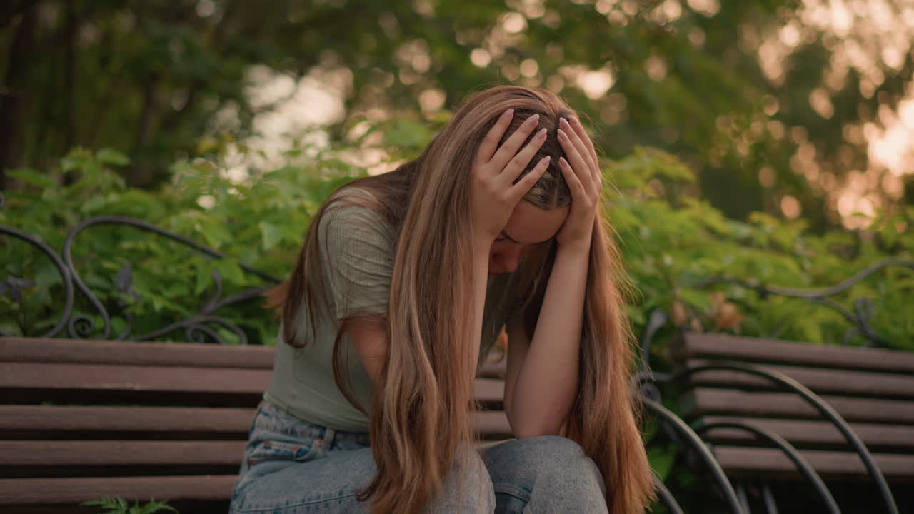 donna seduta su una panchina di legno durante l'ora d'oro, china la testa pensierosa, capelli lunghi che scendono in cascata sulle spalle, circondata da lussureggiante verde, con la calda luce solare che filtra attraverso le foglie