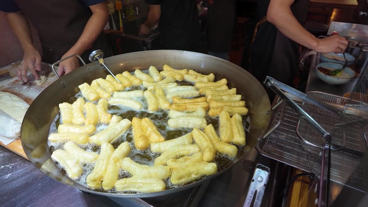 Primo piano di bastoncini di pasta dorati che friggono in olio bollente in un mercato affollato.