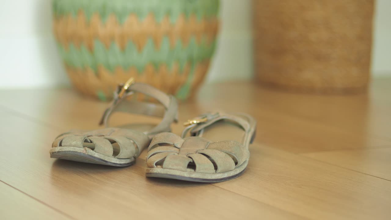 A pair of light-colored sandals on a wooden floor with blurred decorative pots in the background