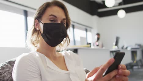 mujer de negocios caucásica con máscara facial y usando un teléfono inteligente en la oficina