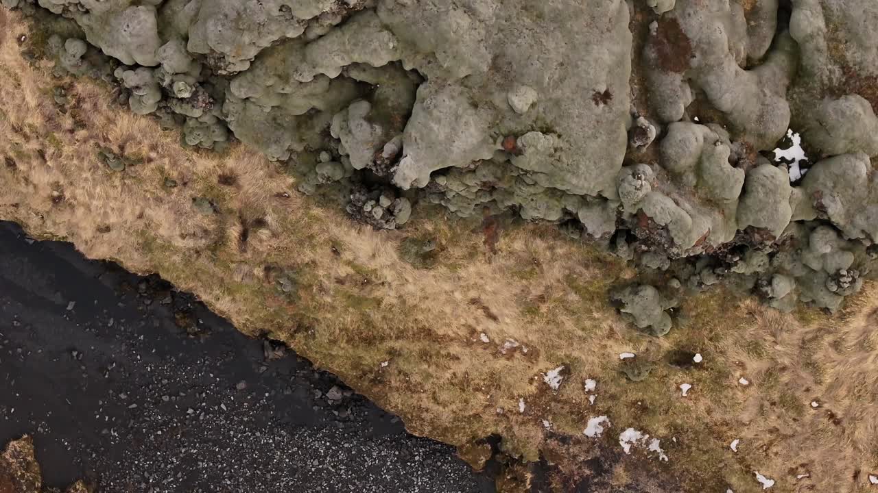 Top-down aerial view of Fjaðrárgljúfur Canyon near Kirkjubæjarklaustur, Iceland, showing black riverbed, moss-covered rocks, and barren terrain in early spring.