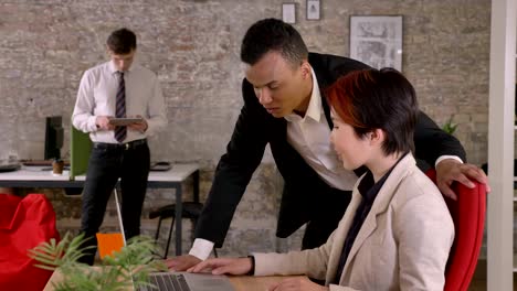 young asian woman and black man talking over laptop in modern office, caucasian man standing in back with tablet