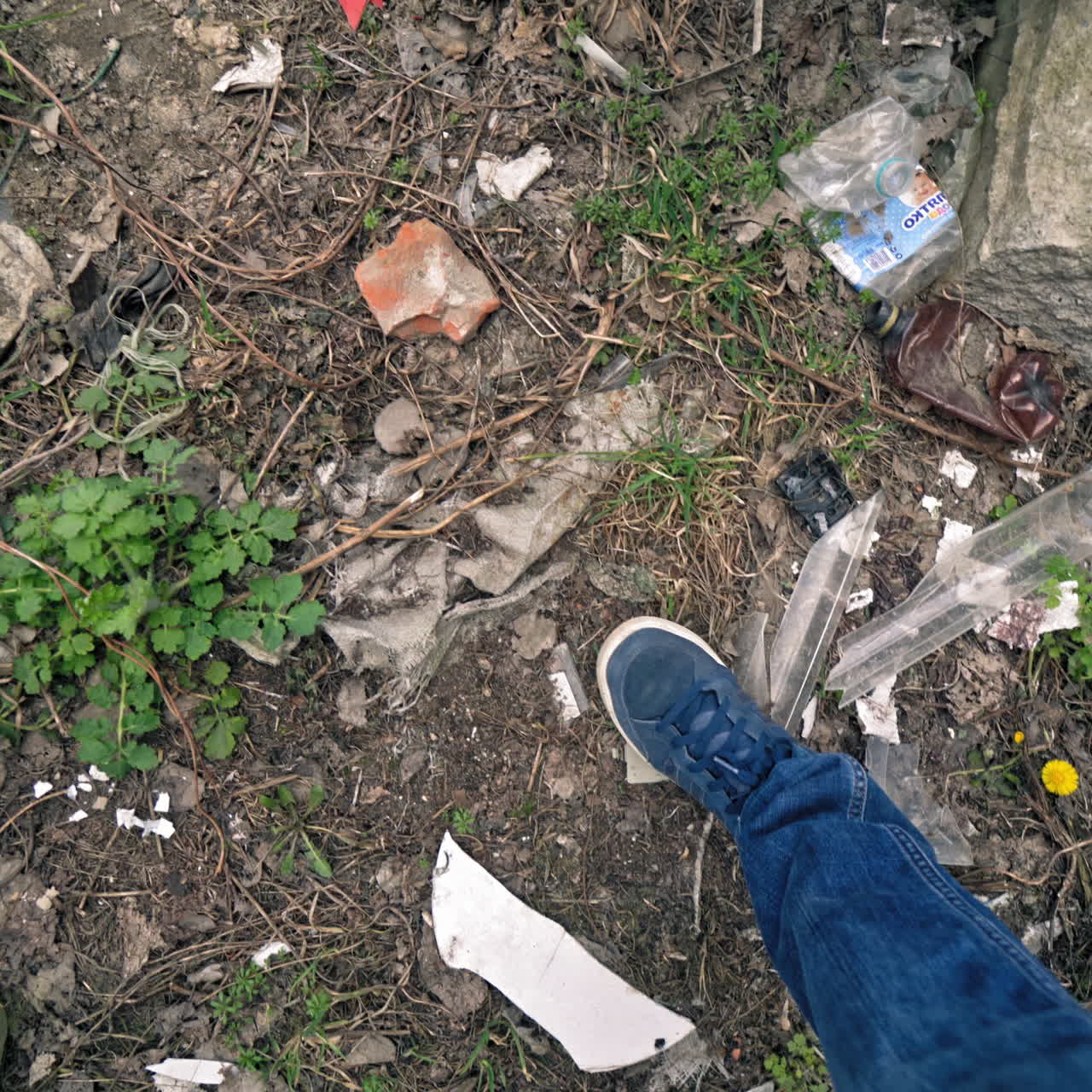 Legs of a man walking through the dirty ground with different garbage outdoors. Male's feet stepping along the dirty place with glass bottles and rubbish.