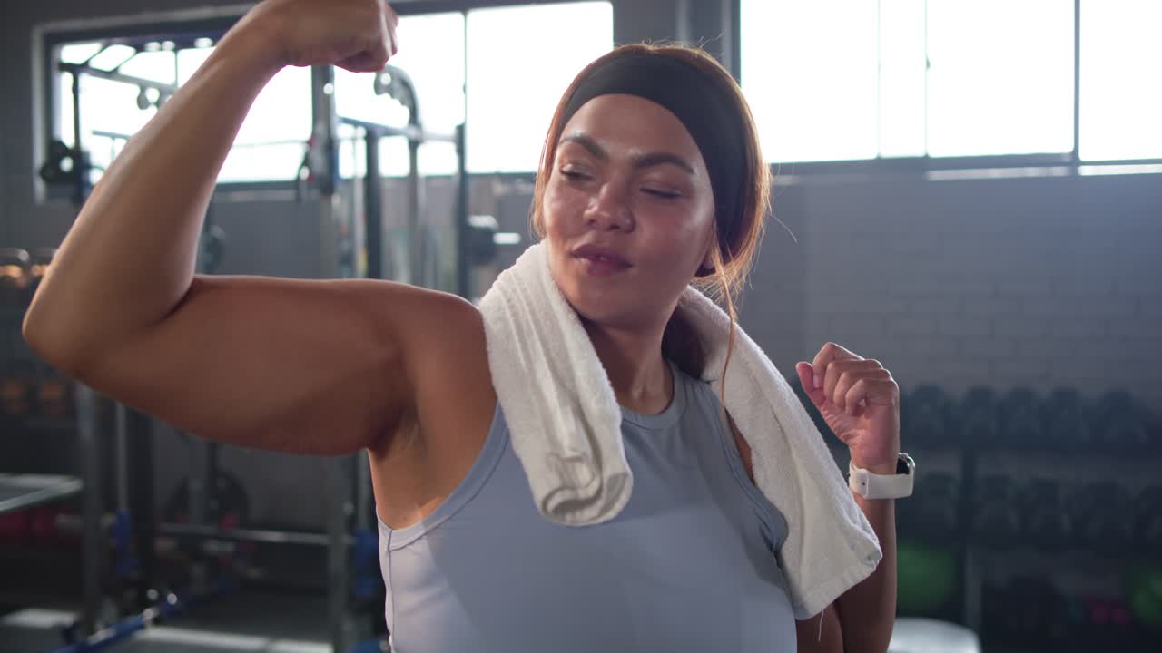 Woman in sleeveless top holding towel behind neck, flexing bicep in gym, checking results on watch