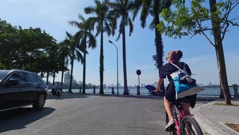 POV-Following-Behind-Female-Tourist-Cycling-Beside-Tay-Ho-Lake-On-Sunny-Day-In-Hanoi