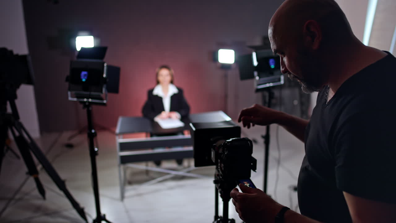 Bald, a bit overweight cameraman arranges a camera on tripod. Woman sits at desk at blurred backdrop in the light of soffits. Blogging concept.