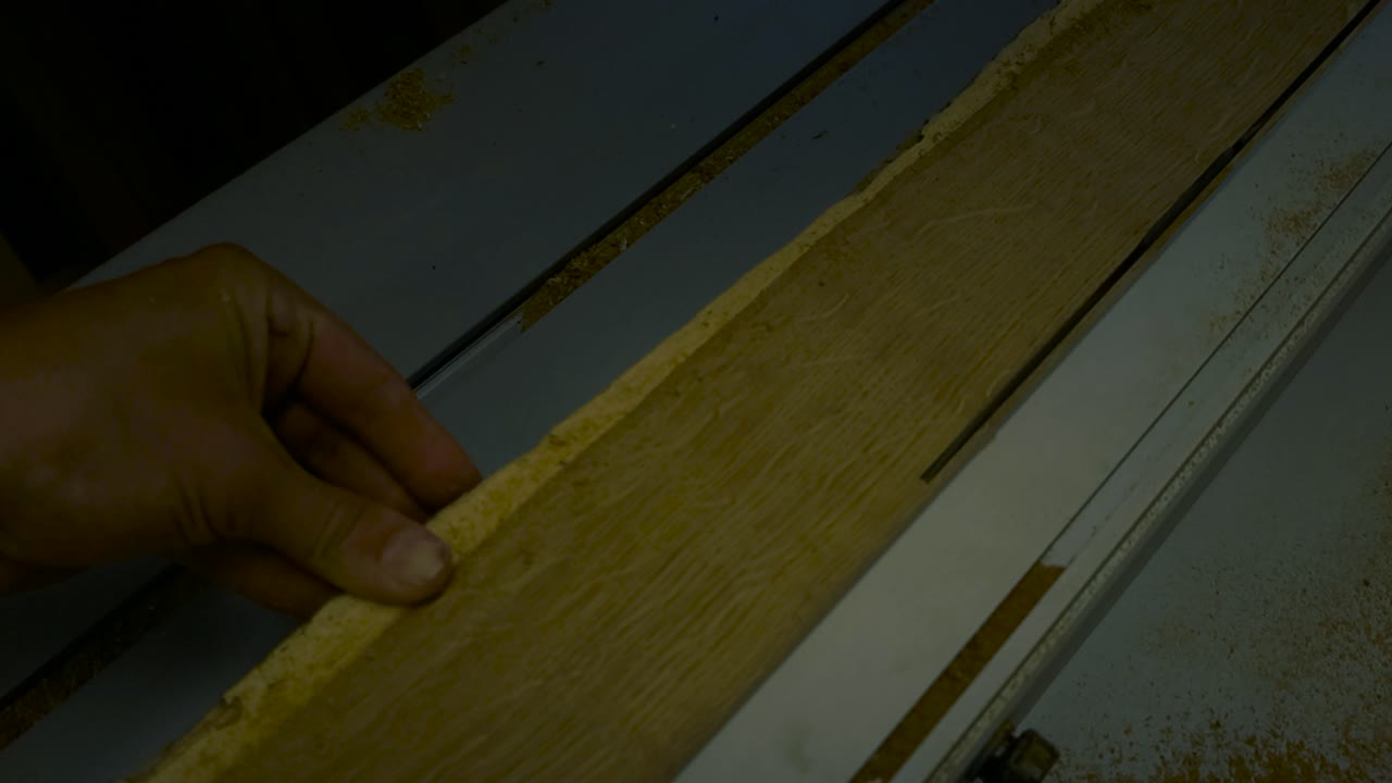 Primo piano cinematico della mano di un artigiano che spinge una lastra di quercia dorata attraverso la lama di una sega da banco, con segatura che vola nell'officina.