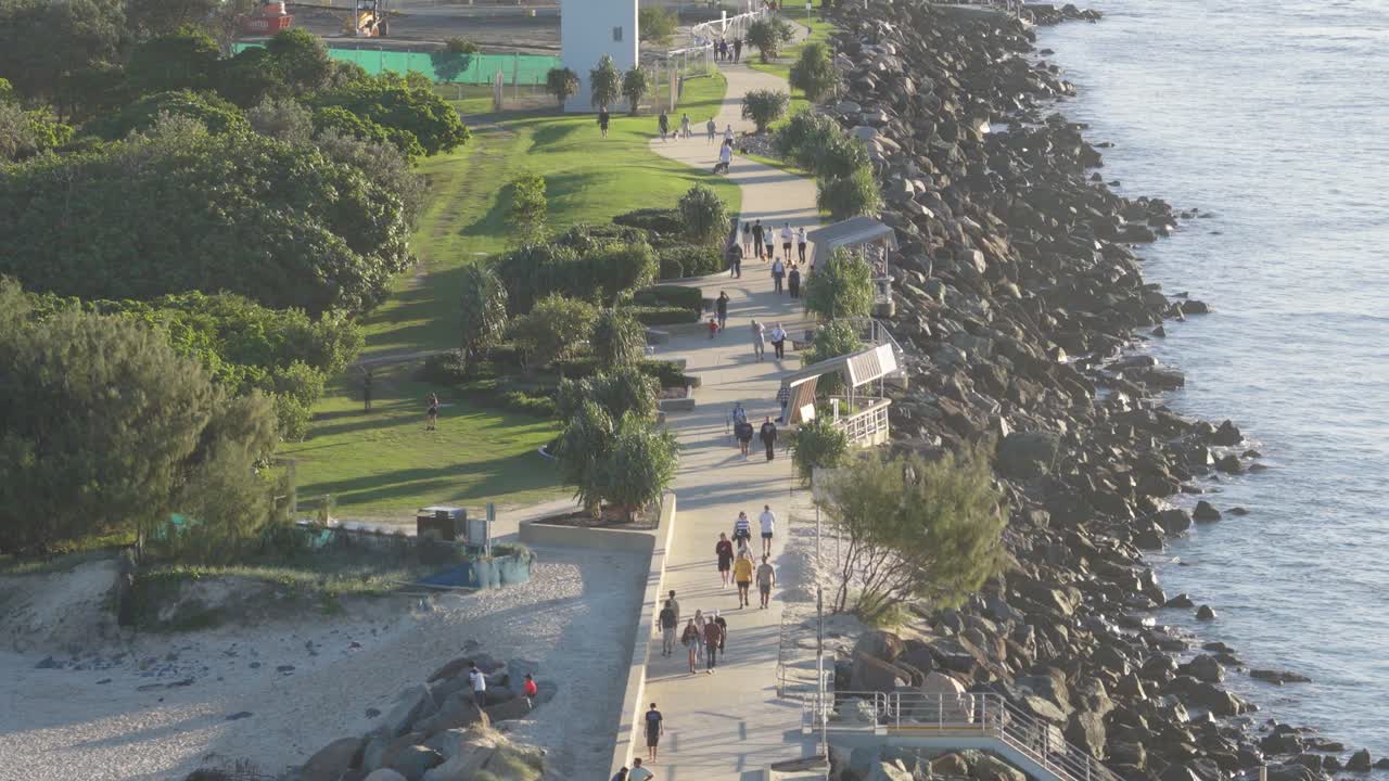Drone captures people strolling on a scenic seaside path with lush greenery and ocean views