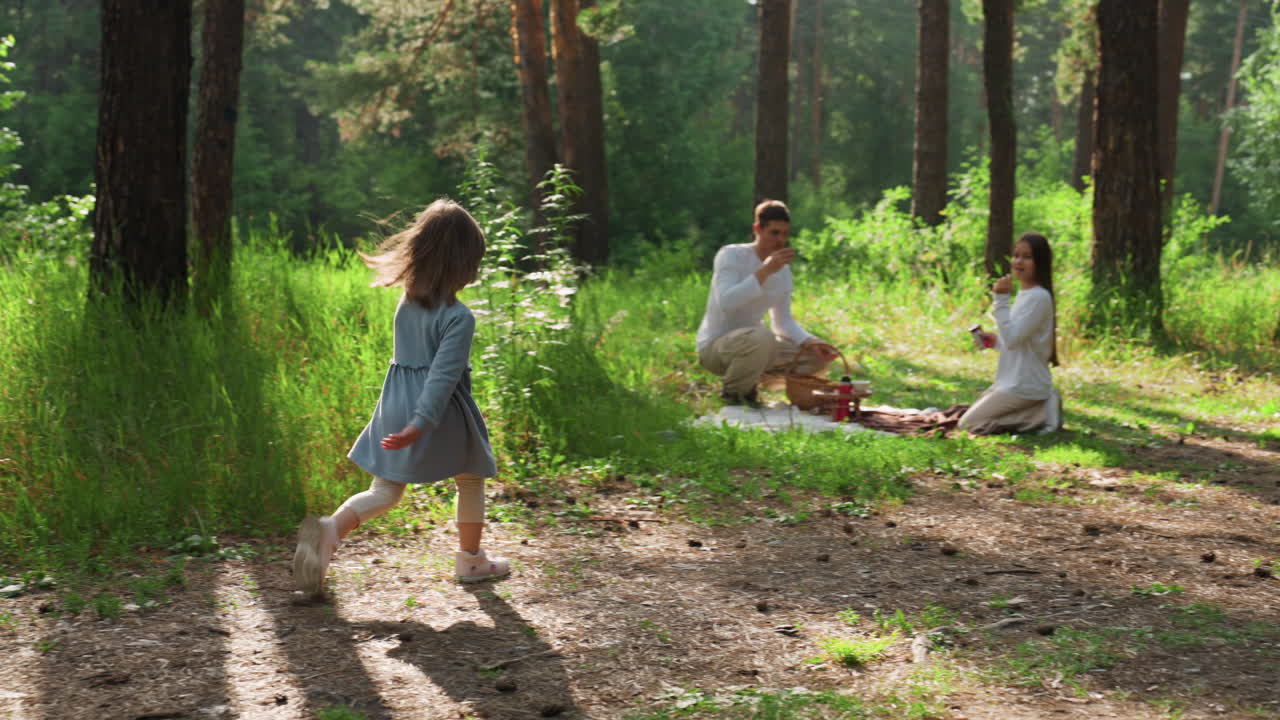 Tiny explorer confidently walking through sunlit forest toward siblings seated on picnic mat surrounded by greenery, capturing warmth, and childhood adventure in peaceful outdoor family setting