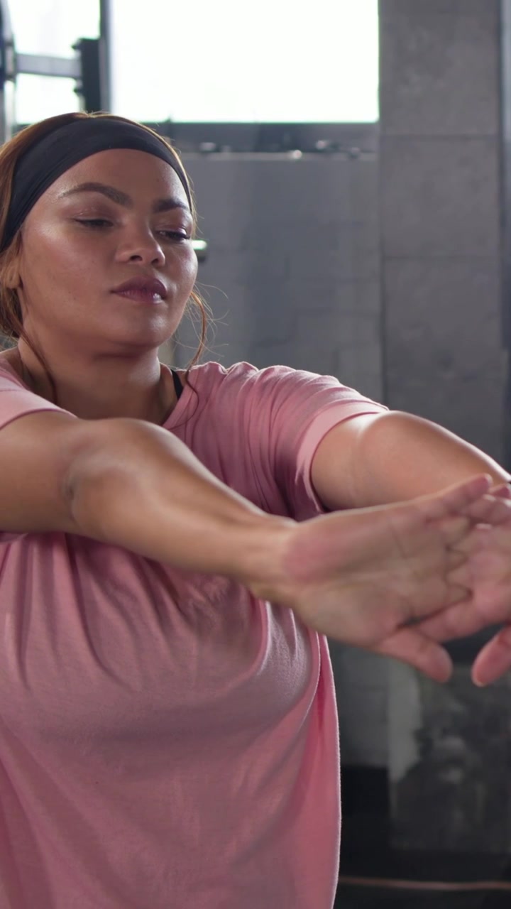 Video verticale: Donna in maglietta rosa si allunga a sinistra, facendo torsioni in palestra e riscaldando le spalle vicino a una rastrelliera.