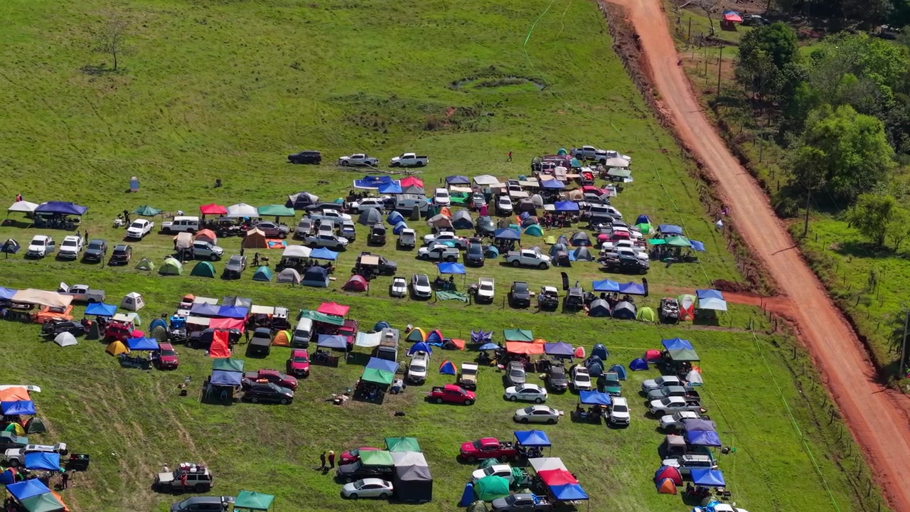 Video drone cattura un vivace campeggio del WRC Rally 2026 a Nueva Alborada, Paraguay, con tende colorate, auto parcheggiate, campi erbosi, strada sterrata rossa e un'atmosfera festosa di folla all'aperto.