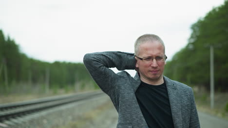 close-up view of a man in glasses and a gray jacket standing on a railway track, looking distressed as he rubs his head with one hand and touching the back of his head