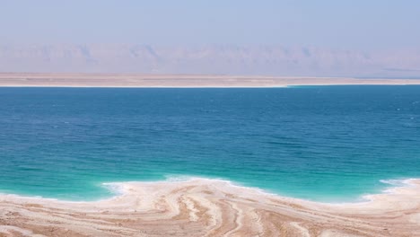 Vista-Panorámica-Del-Mar-Muerto-Con-Agua-Salina-Turquesa-Y-Depósitos-De-Sal-Con-Costa-Blanca-De-Arenas-Movedizas-En-El-Valle-Del-Rift-En-Jordania,-Oriente-Medio