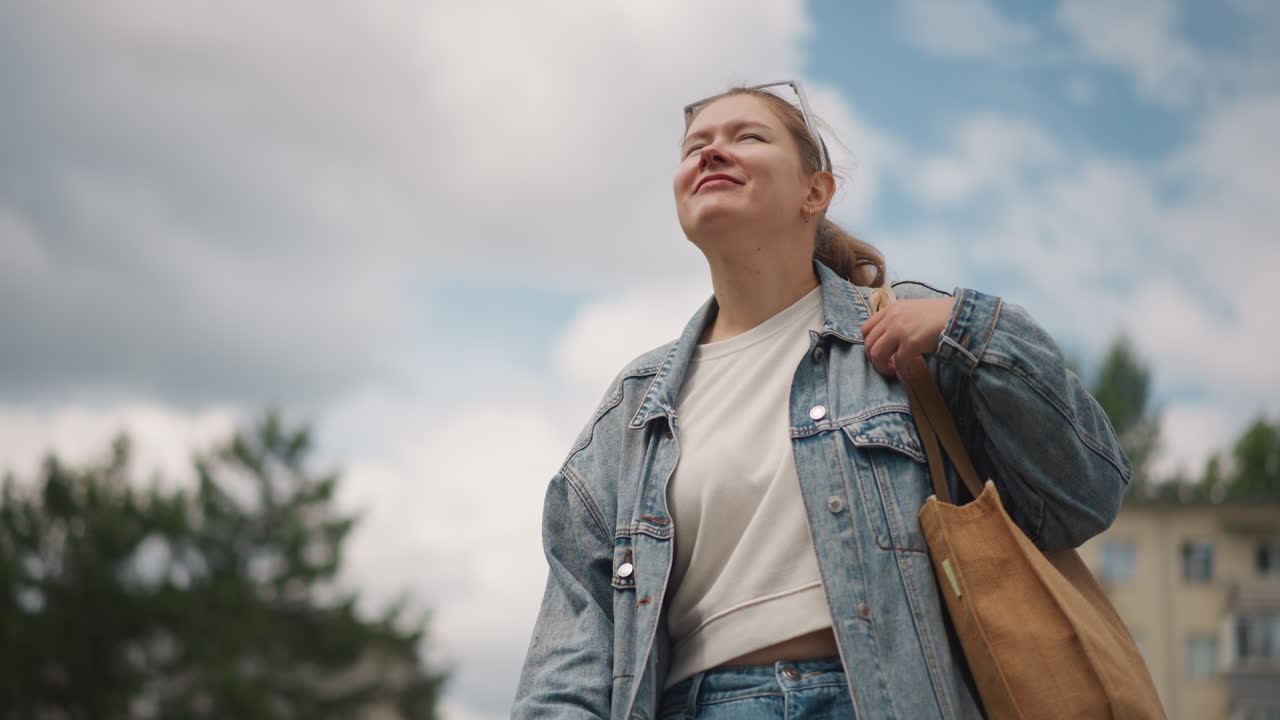 Donna bianca seduta su una panchina che guarda il cielo, con giacca di jeans e tote bag. Ruoli di studentessa e viaggiatrice rilassata e sognante, con sfondo di un appartamento suburbano. Nuvole soffici e luce solare, sguardo verso l'alto, svago all'aria aperta.