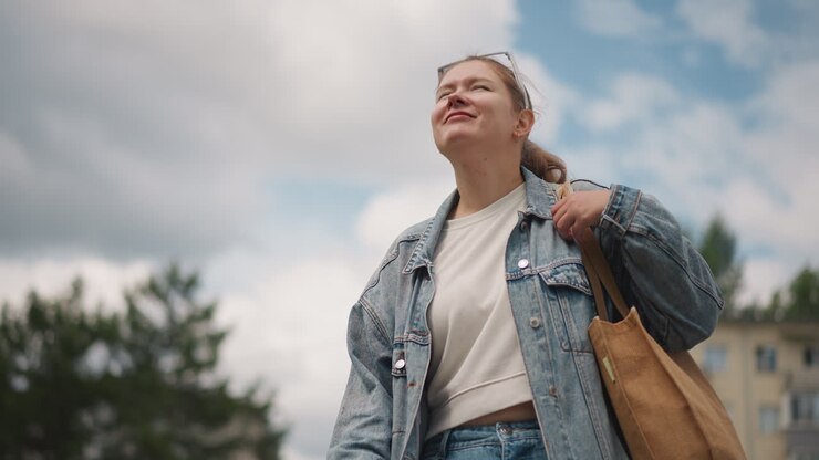 Mujer blanca sentada en un banco mirando al cielo, con chaqueta de mezclilla y bolso tote. Roles de estudiante y viajera relajada y soñadora. Fondo de apartamento suburbano. Nubes suaves y luz solar. Mirada hacia arriba. Ocio al aire libre.