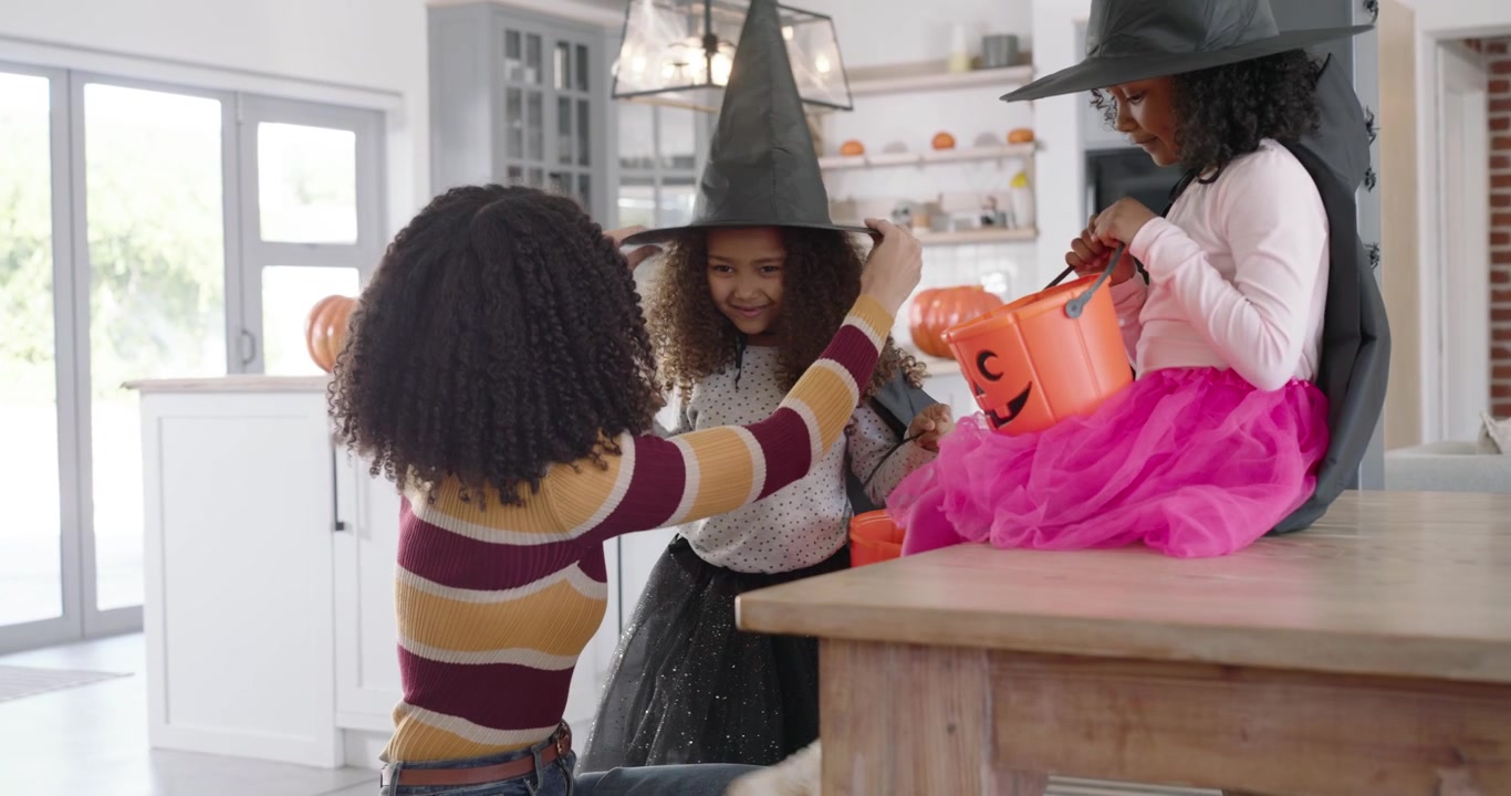 African American mom kneeling in kitchen, placing witch hats as girls arriving holding pumpkin pail