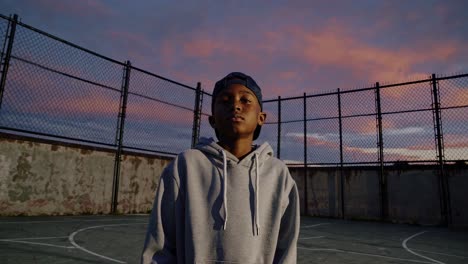 a low-angle video shot of a person in a hoodie on a basketball court at sunset, with a dramatic sky