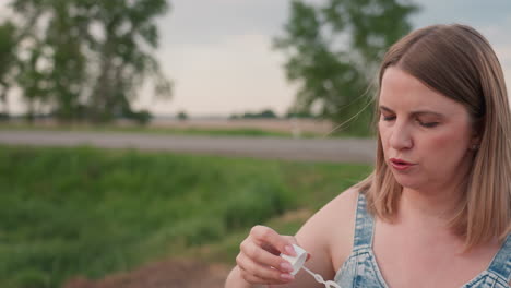 side profile young woman in denim dress gently blowing translucent bubbles toward open roadside field, subtle bubbles drifting in soft breeze against blurred trees and parked car