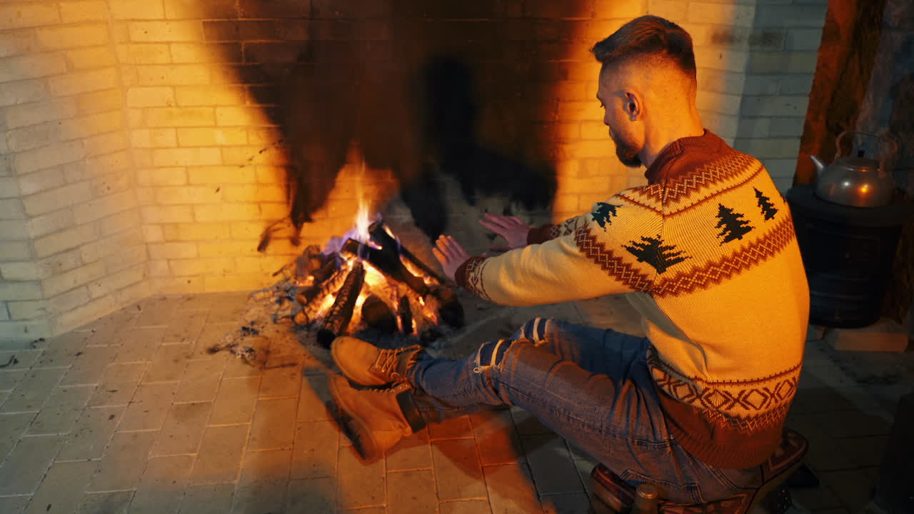 Man is sitting near the fireplace. Young male is warming hands near the fire inside. Cozy evening by the fireplace at home. Cold season concept.