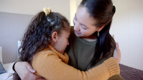 Happy-biracial-mother-comforting-and-embracing-daughter-on-bed-in-sunny-bedroom