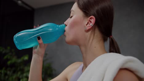 Side-view-close-up-of-a-confident-brunette-girl-in-a-purple-sports-top-with-a-white-towel-on-her-shoulder-drinks-water-from-a-blue-sports-bottle-in-a-modern-apartment-after-playing-sports-in-the-evening