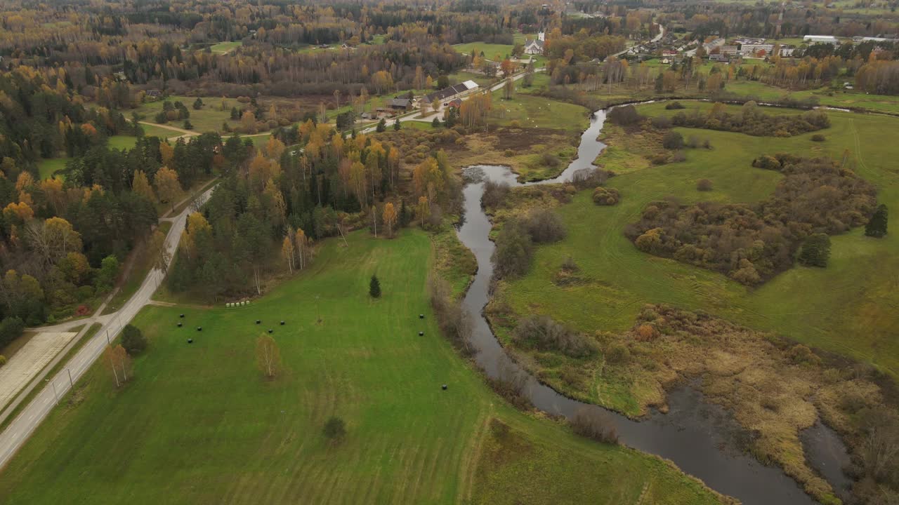 A drone captures the small town of Jaunpiebalga in summer, showcasing its peaceful streets, rooftops, and lush green surroundings under the bright sunlight.