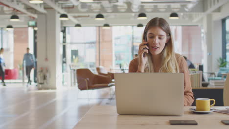 Businesswoman-Sitting-At-Desk-On-Phone-Call-In-Modern-Open-Plan-Office-With-Colleagues-In-Background