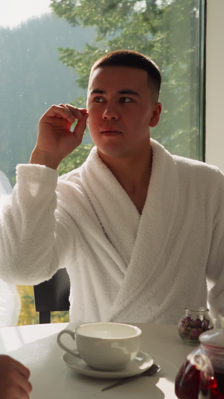 남자가 테이블에 앉아 있는 여자 에 있는 컵에 차 꽃잎을 떨어뜨리고, 남자는 아침에 함께 아침식사하는 뜨거운 차를 즐기며 재미있는 순간을 만니다.