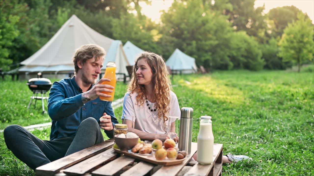 Et par spiser morgenmad i naturen ved glamping. Drikkevarer, mad, snak og smil. Telt i baggrunden. Slow motion