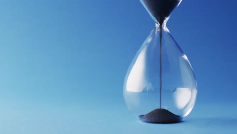 Video-of-close-up-of-hourglass-with-black-sand-pouring-with-copy-space-on-blue-background