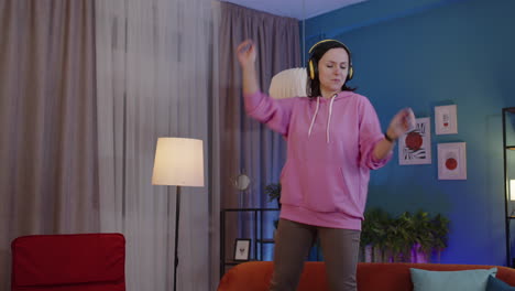 Happy-adult-woman-in-headphones-dancing-and-jumping-while-listening-music-at-home-alone-on-sofa