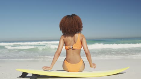 Rear-view-of-African-American-woman-sitting-on-her-surfboard