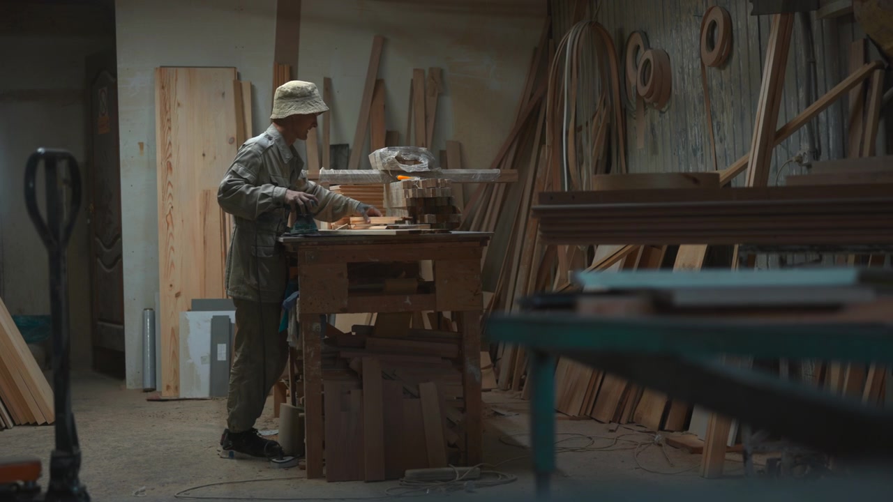 Woodworker in dim workshop reaches across cluttered workbench, aligning stacked cut boards, steadying timber pieces, holding palm sander, with sawdust, tools, muted light, careful inspection and